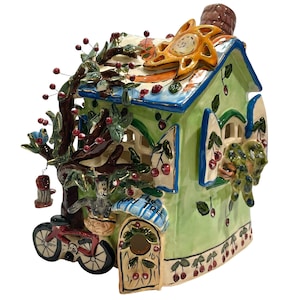 Puede incluir: Una escultura de cerámica con forma de casa en verde claro con techo azul, que presenta un árbol marrón con bayas rojas, una bicicleta pequeña, un adorno de sol y una pajarera. La puerta tiene un toldo azul con el texto The Cherry Tree impreso.