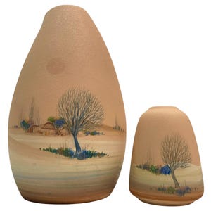 May include: Two beige ceramic vases with a textured matte finish. Each vase features a hand-painted landscape scene depicting a bare tree, rolling hills, and small colorful bushes. The taller vase is teardrop-shaped, while the shorter is cylindrical.