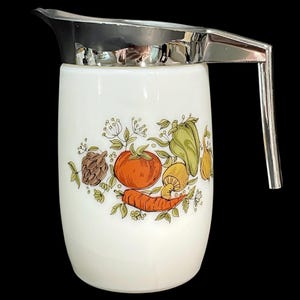 May include: A white milk glass pitcher featuring a chrome lid and handle. The body is decorated with a colourful botanical illustration of a tomato, a carrot, a green pepper, a mushroom, and an onion surrounded by delicate green leafy sprigs.