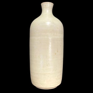 Puede incluir: Una botella alta de cerámica blanquecina con cuello estrecho y cuerpo redondeado. La botella tiene una superficie lisa, ligeramente texturizada con sutiles líneas horizontales y una pequeña base. El fondo es negro.