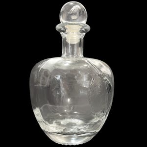 Op de afbeelding: Een heldere glazen karaf met een ronde body en een bolvormige stopper. De karaf heeft geëtste details en een kurkstopper. De karaf wordt waarschijnlijk gebruikt voor het serveren van dranken. Hoogte ongeveer 20 cm.