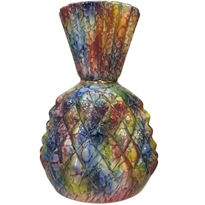Puede incluir: Un jarrón de cerámica con forma de piña y borde acampanado. La superficie presenta un esmalte jaspeado multicolor en tonos azul, amarillo, rojo y morado, acentuado por un patrón de rejilla de diamantes en relieve que crea un efecto texturizado.