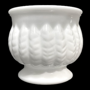 May include: A white, opaque glass planter with a decorative, textured pattern. The planter has a rounded body, a flared rim, and a pedestal base. The design features vertical, feather-like ridges around the body of the planter.