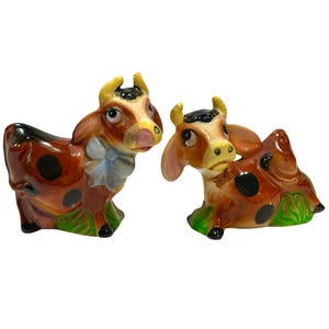 May include: Two ceramic cow figurines featuring brown and white spotted patterns, yellow horns, and green grass bases. One cow stands upright with a light blue bow around its neck, while the other cow is depicted in a reclining, resting position.