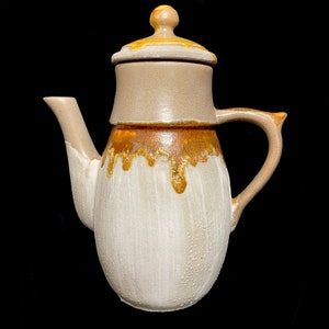 Puede incluir: Una tetera de cerámica con un esmalte blanco y un esmalte marrón con goteo. La tetera tiene un cuerpo redondeado y una tapa con un pomo.