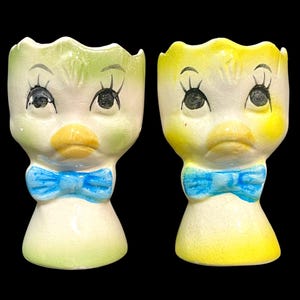 May include: Two ceramic egg cups shaped like stylized bird heads with scalloped rims. One cup features green accents, while the other has yellow accents. Both have painted black eyes with long eyelashes, orange beaks, and bright blue bow ties.