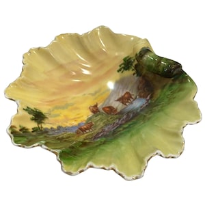 May include: A scalloped, shell-shaped ceramic dish featuring a hand-painted pastoral scene of brown cattle grazing in a green field under a yellow and orange sunset sky. The dish has a glossy finish with a dark green, rolled handle detail.