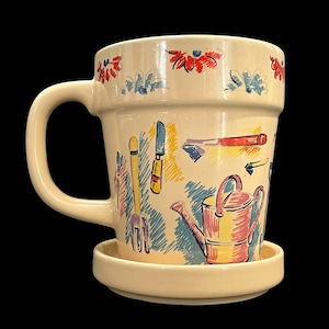 May include: Cream-colored ceramic mug with a matching saucer. The mug features a wraparound illustration of gardening tools and a watering can in pastel colors. Red and blue floral accents adorn the rim. The mug has a large, curved handle.