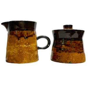 May include: A brown and black ceramic creamer and sugar set with a textured glaze. The creamer has a handle and the sugar bowl has a lid.