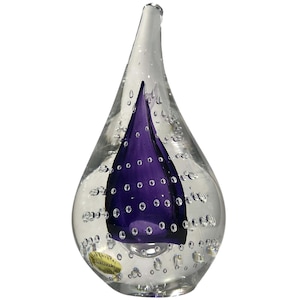 May include: A clear, teardrop-shaped glass sculpture featuring a solid, deep purple core encased within the transparent glass. Numerous small, spherical air bubbles are suspended throughout the clear glass body, and a small gold sticker is attached.