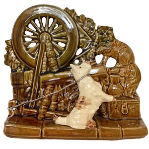 May include: A ceramic figurine depicting a dog pulling a cart with a spinning wheel. The figurine is primarily brown and cream-colored, with a detailed design. The dog is cream-colored with brown spots. The spinning wheel is brown.