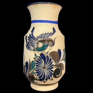 May include: A beige ceramic vase with a blue rim and a blue stripe around the neck. The vase features a hand-painted design of a bird with blue and teal wings, perched above blue floral elements and brown accents.