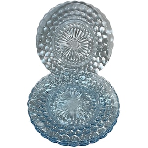 Può includere: Due piatti di vetro blu chiaro con un motivo strutturato a bolle sui bordi. Il centro di ogni piatto presenta un disegno a stella. I piatti sono impilati, mostrando i loro dettagli intricati e il design elegante.