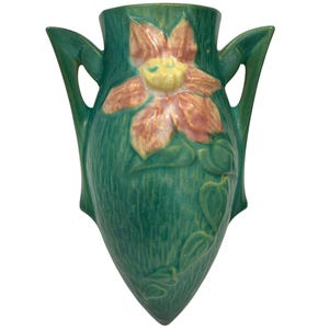 Puede incluir: Un florero de pared de cerámica verde con un diseño en relieve de una flor grande de color rosa con centro amarillo. El florero tiene dos asas laterales y una superficie texturizada con detalles de enredaderas y hojas en el cuerpo frontal.