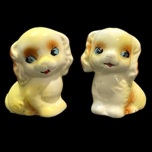 May include: Two ceramic puppy figurines. The puppies are sitting, with yellow and white bodies, brown spots, blue eyes, and red mouths. The figurines have a glossy finish and are set against a black background.