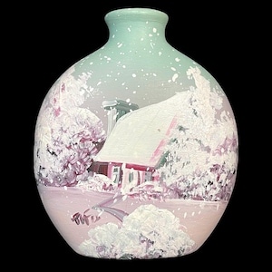 May include: A round ceramic vase with a winter scene painted on it. The vase is light green with white speckles and a pink base. The scene depicts a snow-covered house with pink and red accents, surrounded by trees and a snowy landscape.