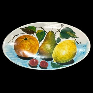 Puede incluir: Una bandeja de servicio ovalada de cerámica con un bodegón pintado a mano de una naranja grande, una pera verde, un limón amarillo y dos cerezas rojas pequeñas. Las frutas están representadas con hojas verdes y tallos marrones sobre un fondo azul.