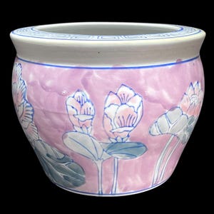May include: A round, pink ceramic planter with a white rim and blue accents. The planter features a floral design with pink and blue lotus flowers and leaves. The rim has a blue geometric pattern.