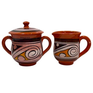 May include: A ceramic sugar bowl with a lid and a matching creamer pitcher. Both pieces are orange with brown, white, and yellow hand-painted geometric swirl patterns. The sugar bowl features two handles and the word PANAMA printed on the front.
