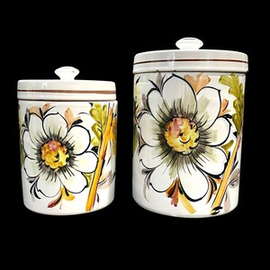 May include: Two white ceramic canisters with lids, each featuring a hand-painted floral design with large white petals, yellow centres, and green and orange foliage accents. The cylindrical containers have a brown horizontal line near the top rim.