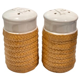 May include: A pair of ceramic salt and pepper shakers featuring a white glazed top with embossed S and P letters and a textured, golden-tan woven basket-weave pattern on the lower body. Each shaker has small holes on the top for dispensing.