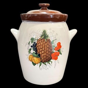 Puede incluir: Un tarro de galletas de cerámica vintage con tapa y asas marrones. El tarro es de color blanquecino con un diseño de frutas colorido que presenta una piña, cerezas y otras frutas. Un artículo de cocina clásico.