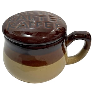 May include: A brown ceramic coffee mug with a lid. The mug features a gradient of colours, from light tan at the base to dark brown at the top. The lid is embossed with the words "ALTE KAFFE". It has a curved handle.