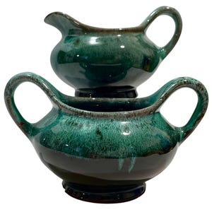 Puede incluir: Juego de servir de cerámica de dos piezas en un esmalte verde intenso con ribete marrón oscuro. El juego incluye una pequeña jarra de crema con pico y asa, apilada sobre un cuenco más grande con dos asas. Las piezas tienen un acabado brillante.