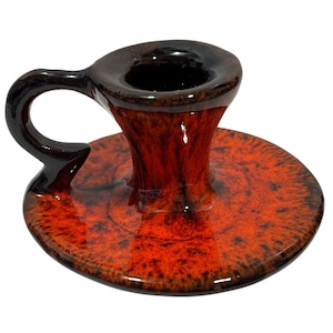 Puede incluir: Un candelero de cerámica con asa. El candelero está esmaltado en tonos rojos, marrones y negros. La base es redonda y el asa se curva con gracia. El interior del candelero es negro.