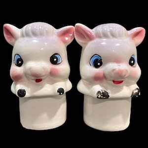 May include: A pair of ceramic salt and pepper shakers shaped like pig heads with white bodies, pink ears and cheeks, blue eyes, and red mouths. The figures feature small black hooves resting on the cylindrical base with a visible crackle glaze finish.
