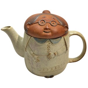 May include: A ceramic teapot shaped like a person with a brown head wearing round glasses as the lid. The body is a speckled cream colour with a blue collar, featuring embossed hands, a buttoned front, and a patterned hem. It has a curved handle and spout.