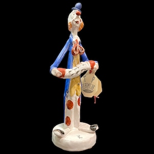 May include: A tall ceramic clown figurine wearing a blue coat, yellow vest, and white pants with orange polka dots. The clown has a blue hat, red nose, and holds a white accordion. A beige tag with the text TP CERAMICHE hangs from the instrument.