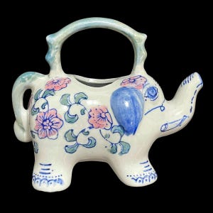 May include: A white ceramic teapot shaped like an elephant with blue and pink floral designs. The teapot has a handle and a spout.