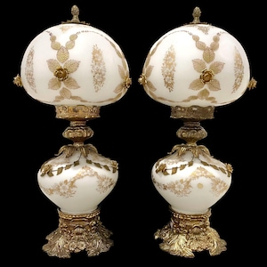 Può includere: Coppia di lampade da tavolo antiche. Ogni lampada presenta un paralume in vetro bianco con disegni floreali e a foglie dorate. La base e gli accenti sono in metallo dorato ornato. Le lampade hanno un'estetica classica e vintage.