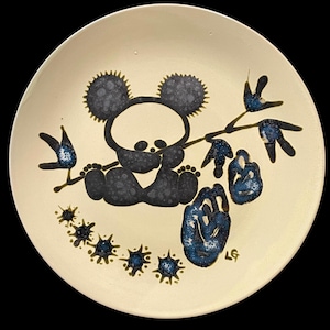 May include: A white ceramic plate featuring a black silhouette of a koala bear holding a branch with blue and green leaves. The plate is decorated with black and blue splatter paint.