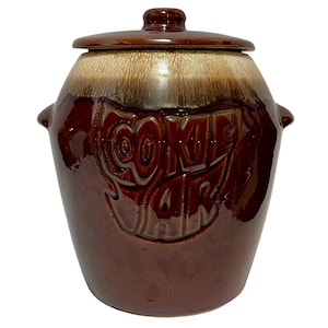 Può includere: Un barattolo per biscotti in ceramica marrone con finitura lucida, dotato di coperchio rimovibile e due manici laterali. La parte anteriore presenta le parole COOKIE JAR in rilievo con grandi lettere maiuscole e uno smalto color crema.