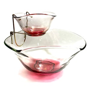 Puede incluir: Un juego vintage de vidrio para chips y salsa con un tazón transparente y un tazón rosa más pequeño en la parte superior. El juego tiene un soporte de metal dorado que une los dos tazones.