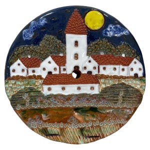 Puede incluir: Un colgante de pared de cerámica que representa una escena de pueblo con casas blancas con techos rojos bajo una luna llena. Las casas están rodeadas de árboles verdes y un muro de piedra. El fondo es un cielo azul oscuro con nubes blancas.