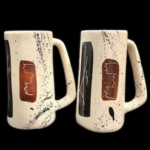 Op de afbeelding: Twee crèmekleurige keramische mokken met zwarte en bruine abstracte ontwerpen. De mokken hebben grote handvatten en zijn versierd met zwarte verfspatten. Elke mok heeft een rechthoekige bruine vorm met een witte lijntekening.