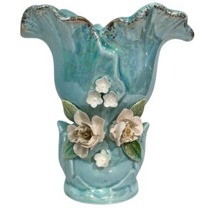 Peut inclure: Un vase en céramique bleu clair irisé avec un bord ondulé bordé d'or. Le corps présente des appliques florales blanches en trois dimensions avec des feuilles vertes et de petites fleurs blanches fixées à l'avant, créant un aspect texturé.