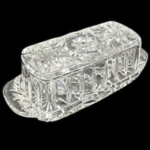 May include: A clear, rectangular glass butter dish with a lid. The dish features a diamond-cut pattern on the lid and sides, and a scalloped edge on the base. The butter dish is designed to hold a standard stick of butter.