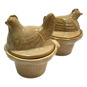 Può includere: Due contenitori in ceramica color marrone chiaro con coperchio, ciascuno caratterizzato da una statuetta di gallina scolpita sul coperchio. La base di ogni contenitore presenta un motivo a griglia che ricorda un cesto intrecciato.