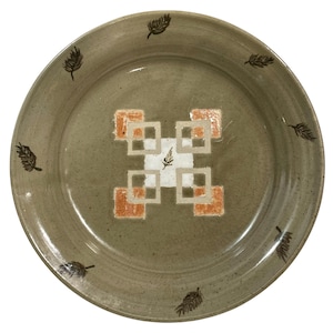 May include: A round ceramic plate with a speckled, olive-green glaze. The centre features a geometric design of white squares and orange rectangles, with a small fern-like detail. Dark brown leaf motifs are evenly spaced around the rim.