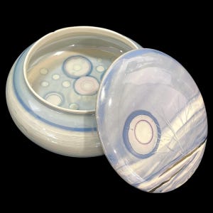 May include: A ceramic bowl and lid in shades of light blue and white. The bowl is round with blue stripes, and the lid is oval-shaped. The design includes circular patterns in blue, white, and pink, creating a decorative effect.