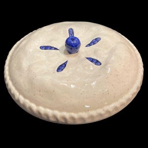 Puede incluir: Una fuente para pastel de cerámica con un esmalte color crema y un borde decorativo ondulado. La tapa de la fuente para pastel presenta un pomo azul en forma de arándano y recortes azules en forma de hoja. La fuente para pastel está sobre un fondo negro.