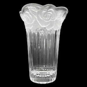May include: A clear glass vase with a fluted body and a scalloped rim. The rim features frosted rose and leaf designs. The vase is transparent, allowing light to pass through, and is set against a black background.