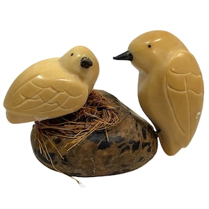May include: Two carved stone birds, one perched on a nest. The birds are a light tan colour with black beaks and eyes. The nest is a mottled brown and green colour, with strands of straw.