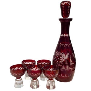 Può includere: Un decanter in vetro rosso intenso con tappo abbinato e cinque piccoli bicchieri coordinati. Ogni pezzo presenta intricati disegni incisi in trasparente raffiguranti palme e strutture architettoniche, su basi in vetro esagonale sfaccettato.