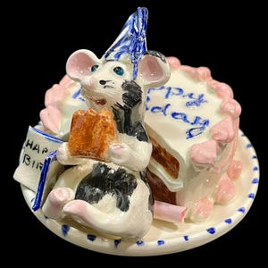 May include: A ceramic figurine of a white and black mouse wearing a blue party hat, holding a brown treat, sitting on a plate next to a slice of cake with pink frosting and the blue text Happy Birthday, plus a small sign reading HAP BIR.