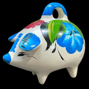 Puede incluir: Una alcancía de cerámica blanca con diseños florales azules, rojos y verdes. La alcancía tiene un asa azul y detalles azules en las orejas y el hocico. Los ojos son negros y las patas blancas.